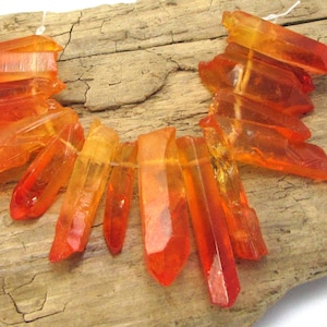 Puede incluir: Un collar de puntas de cristal naranja y rojo, ensartadas con hilo blanco. Los cristales están dispuestos sobre un trozo de madera envejecida, mostrando su color vibrante y formas únicas. Los cristales son una mezcla de translúcido y opaco.