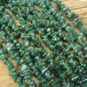 May include: A strand of green gemstone chips, also known as green aventurine chips, arranged in rows on a wooden surface. The chips are small and irregular in shape, with a translucent green color and a slightly sparkly finish.