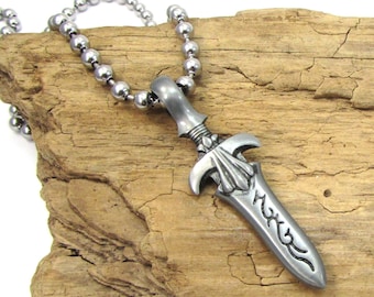 May include: A silver-tone pendant necklace with a dagger-shaped charm. The pendant has detailed carvings and hangs from a ball chain. The necklace is presented on a piece of aged wood.
