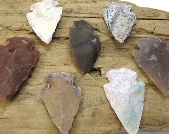 Puede incluir: Una colección de ocho puntas de flecha hechas de varios tipos de piedra, incluyendo verde, blanco, marrón y gris. Las puntas de flecha están dispuestas sobre un trozo de madera a la deriva.