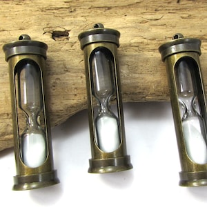 May include: Three antique brass hourglass pendants with clear glass bulbs and white sand. The hourglasses are hanging from a wooden surface.