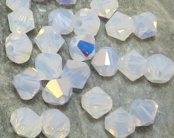 May include: A close-up of a group of iridescent white crystal beads on a gray rock. The beads are faceted and have a rainbow sheen.