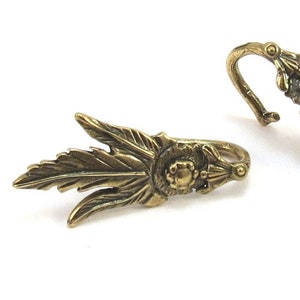 May include: Two gold-toned metal curtain tiebacks with a leaf and floral design. The tiebacks have a hook on one end and a decorative finial on the other.