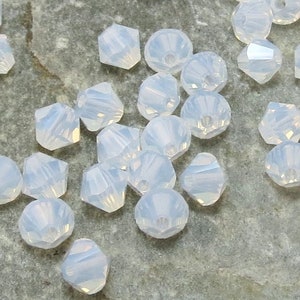 May include: A close-up of a group of small, iridescent white beads on a gray stone surface. The beads are faceted and have a slightly opalescent sheen.