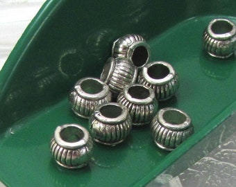 May include: Silver-colored metal beads with a ribbed barrel design. The beads are scattered on a clear, leaf-shaped glass plate and a green scoop. These small beads are suitable for jewelry making and crafting projects.