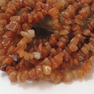 May include: A strand of carnelian chip beads in shades of orange and red. The beads are irregular in shape and size, and they are strung on a clear thread.
