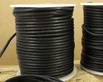 May include: Two spools of black waxed cotton cord, each wound on a white plastic spool. The cord is lying on a piece of driftwood.