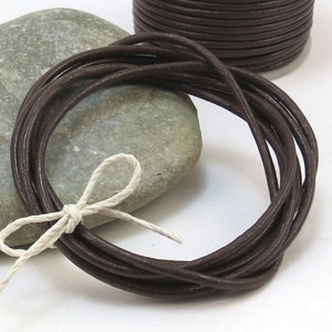 Może przedstawiać: Szpula z ciemnobrązowym skórzanym sznurkiem znajduje się obok wiązki tego samego sznurka, związanego białą nicią. Na zdjęciu znajduje się również szary kamień. Sznurek jest ciasno nawinięty na szpulę i w luźnej spirali.