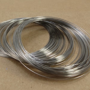 May include: A collection of silver-colored wire coils, neatly arranged in a circular pattern. The thin, metallic wires are tightly wound, creating a dense, textured appearance. The coils are likely used for jewelry making or other craft projects.
