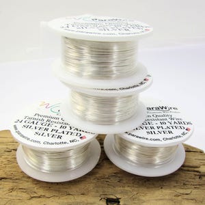 Op de afbeelding: Vier spoelen verzilverde draad zijn op een houten oppervlak gestapeld. Elke spoel is wit met zwarte tekst met "ParaWire" en "24 Gauge - 9,14 meter". De draad is strak opgerold en glanzend.