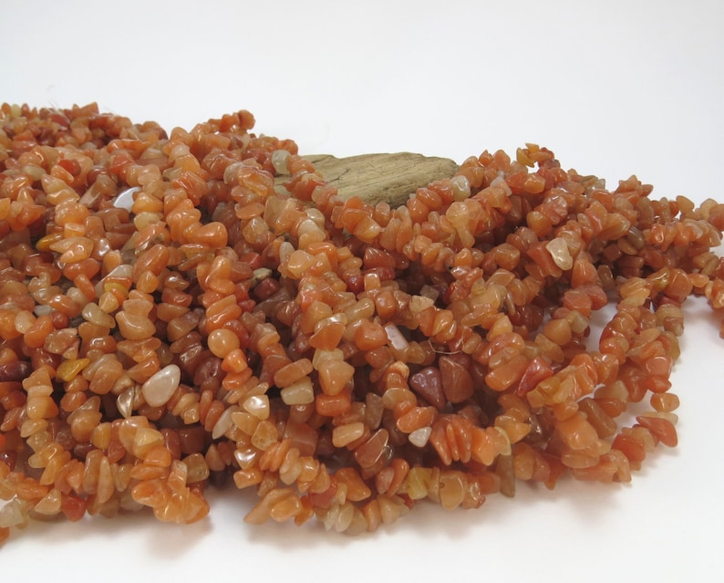 May include: A pile of carnelian chip beads. The beads are a mix of orange and red colors and are irregularly shaped. The beads are arranged in a loose pile on a white surface.
