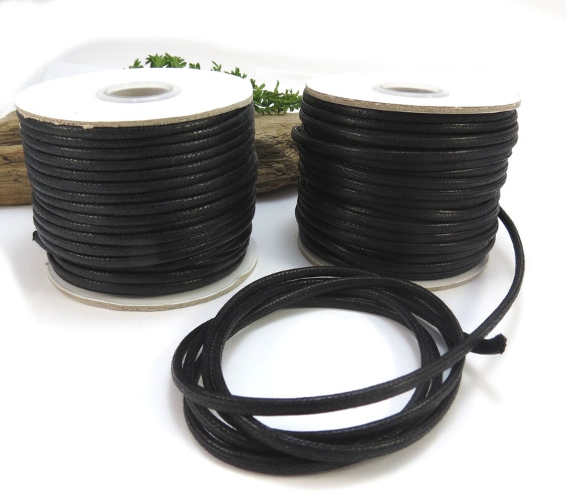 May include: Two spools of black waxed cotton cord, each wound on a white cardboard spool. A length of cord is also shown lying on a white surface.
