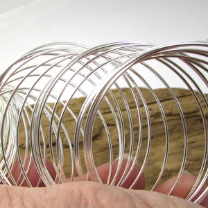 May include: A collection of silver-colored wire bangles, arranged in a circular pattern. The thin, shiny metal reflects light, creating a bright and elegant appearance. The bangles are stacked together, showcasing their delicate design. The background is a piece of driftwood.