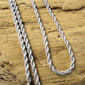K&ouml;nnte beinhalten: Eine silberfarbene Seilkette. Die Kette besteht aus verdrehten Metallgliedern, die ein seilartiges Aussehen erzeugen. Die Halskette wird vor einem strukturierten, hellbraunen Holzuntergrund pr&auml;sentiert.