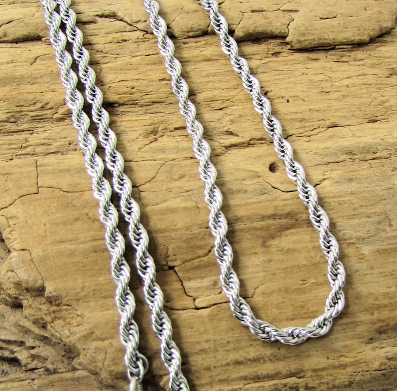 K&ouml;nnte beinhalten: Eine silberfarbene Seilkette. Die Kette besteht aus verdrehten Metallgliedern, die ein seilartiges Aussehen erzeugen. Die Halskette wird vor einem strukturierten, hellbraunen Holzuntergrund pr&auml;sentiert.