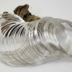 May include: A collection of silver-colored wire coils arranged in a flowing, circular pattern. The coils are set against a white background, with a piece of driftwood in the background. The wire appears to be thin and flexible.