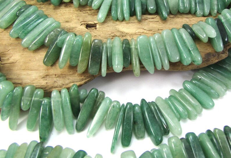 May include: A strand of green jade gemstone beads, each bead is a long, slender, and slightly pointed oval shape. The beads are strung together on a clear thread and are arranged in a staggered pattern.