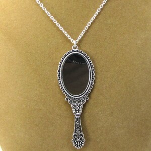 May include: A silver-toned necklace featuring a vintage-style hand mirror pendant. The mirror is oval-shaped and has a detailed frame. The handle of the mirror is ornate and features a heart-shaped design at the bottom.
