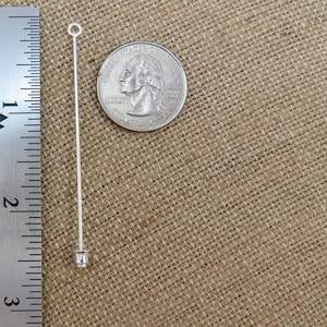 Puede incluir: Un alfiler de cabeza recta plateado con un bucle en la parte superior y un fondo cerrado. El alfiler mide aproximadamente 5 cm de largo y se muestra junto a una moneda de veinticinco centavos de d&oacute;lar estadounidense para comparar el tama&ntilde;o.