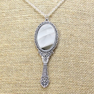 May include: A silver-toned hand mirror necklace with an ornate frame and handle. The mirror is oval-shaped and has a clear reflective surface.