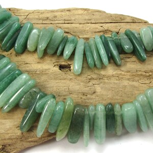 May include: A strand of green jade gemstone beads, each bead is a long, thin, and slightly curved shape. The beads are strung together on a clear thread and are displayed on a piece of driftwood.