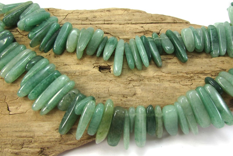 May include: A strand of green jade gemstone beads, each bead is a long, thin, and slightly curved shape. The beads are strung together on a clear thread and are displayed on a piece of driftwood.