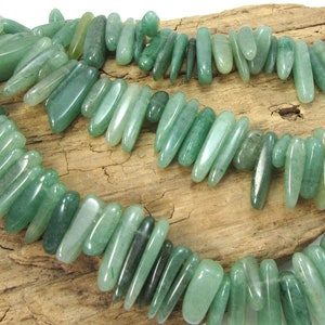 May include: A strand of green jade gemstone beads, each bead is a long, slender, and slightly irregular shape. The beads are arranged in a loose, flowing pattern.
