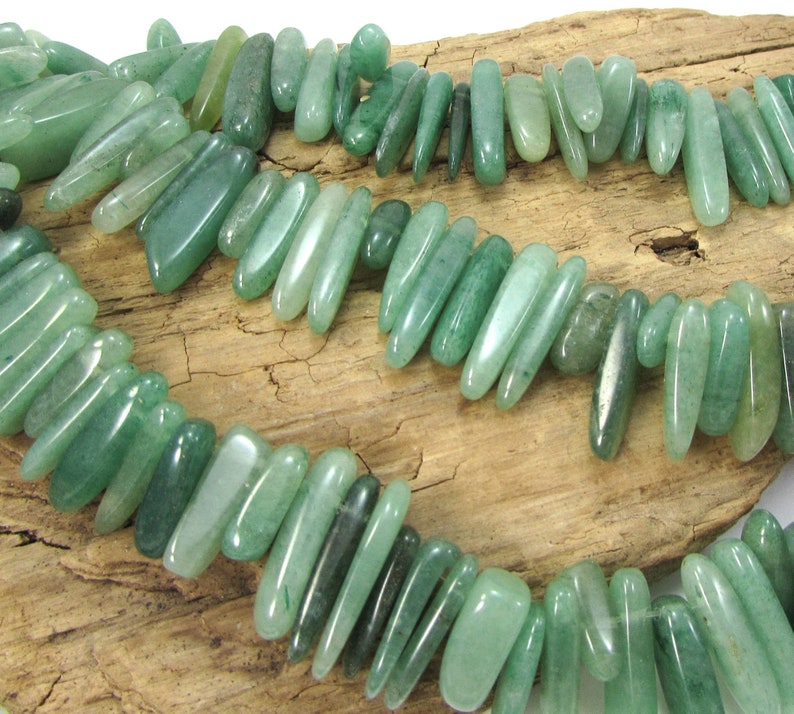 May include: A strand of green jade gemstone beads, each bead is a long, slender, and slightly irregular shape. The beads are arranged in a loose, flowing pattern.