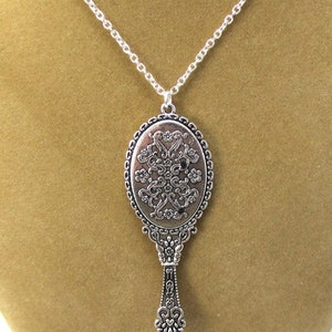 May include: A silver necklace featuring a vintage-style hand mirror pendant. The mirror has an ornate design with floral details.