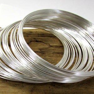 May include: A collection of silver-colored wire bangles, arranged in a circular pattern. The bangles are stacked and intertwined, creating a layered effect. The background is a wooden surface.