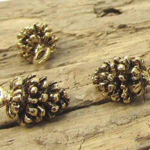 May include: Close-up of several small, gold-coloured pinecone charms. Each charm has a detailed, textured surface and a loop for attaching to jewellery or other crafts. The charms are arranged on a weathered wooden surface.