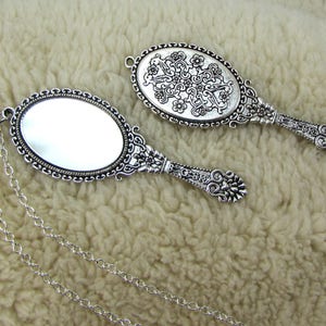 May include: Two silver-toned hand mirrors with ornate designs. One mirror has a plain reflective surface, while the other has a decorative pattern on the back. The mirror with the plain reflective surface is attached to a silver chain.