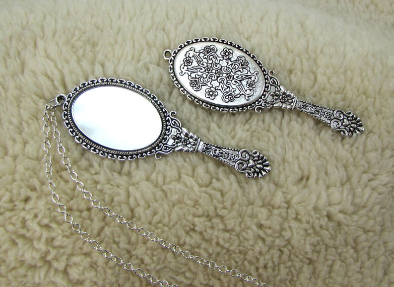 May include: Two silver-toned hand mirrors with ornate designs. One mirror has a plain reflective surface, while the other has a decorative pattern on the back. The mirror with the plain reflective surface is attached to a silver chain.