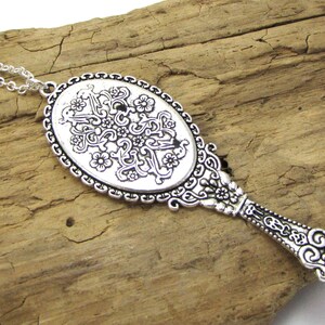 May include: A silver-toned necklace with a decorative mirror pendant. The mirror is oval-shaped and features an intricate floral design. The necklace chain is silver and has a delicate link pattern.