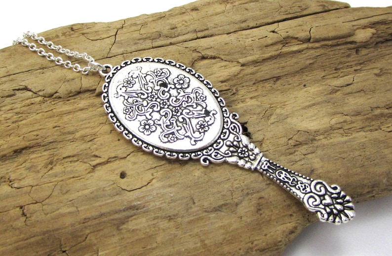 May include: A silver-toned necklace with a decorative mirror pendant. The mirror is oval-shaped and features an intricate floral design. The necklace chain is silver and has a delicate link pattern.