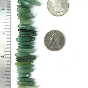 May include: A strand of green jade gemstone beads, each bead is a long, thin, and slightly curved shape. The beads are arranged vertically and are measured against a ruler. The beads are approximately 1 inch in length.
