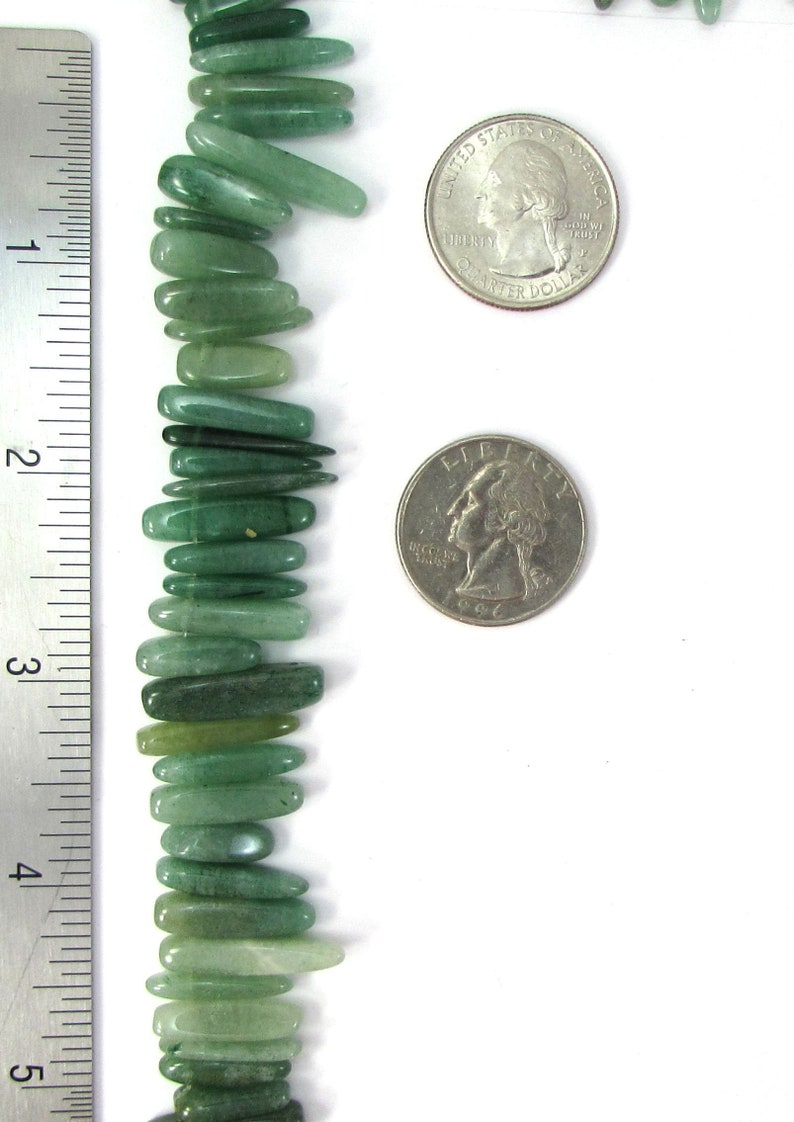 May include: A strand of green jade gemstone beads, each bead is a long, thin, and slightly curved shape. The beads are arranged vertically and are measured against a ruler. The beads are approximately 1 inch in length.