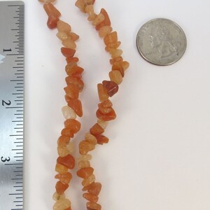 May include: A strand of carnelian gemstone beads, with a mix of light and dark orange shades, is shown next to a ruler and a US quarter for size reference.