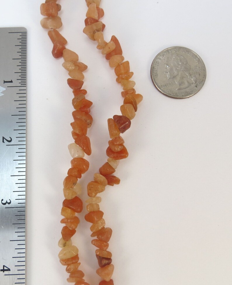 May include: A strand of carnelian gemstone beads, with a mix of light and dark orange shades, is shown next to a ruler and a US quarter for size reference.