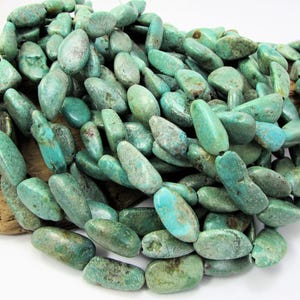 May include: A collection of turquoise gemstone beads in various shades of blue-green. The beads are oval-shaped and appear to be strung together. The beads are resting on a piece of wood.
