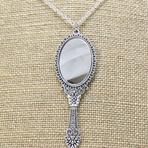 May include: A silver hand mirror necklace with an ornate, detailed frame and handle. The mirror is oval shaped and has a clear reflective surface.
