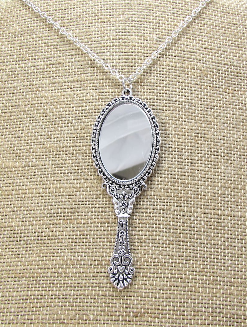May include: A silver hand mirror necklace with an ornate, detailed frame and handle. The mirror is oval shaped and has a clear reflective surface.