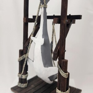 Pu&ograve; includere: Una statuetta di squalo bianco e grigio appesa a una struttura in legno con una corda. La struttura ha una piattaforma in legno ed &egrave; realizzata in legno invecchiato.