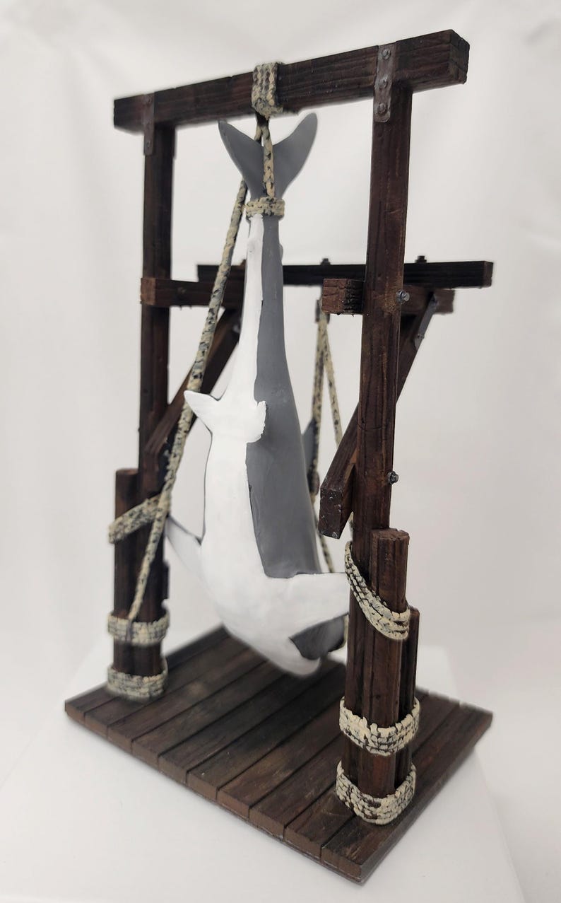 Pu&ograve; includere: Una statuetta di squalo bianco e grigio appesa a una struttura in legno con una corda. La struttura ha una piattaforma in legno ed &egrave; realizzata in legno invecchiato.