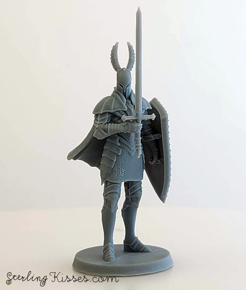 May include: A gray 3D printed miniature of a knight in full armor, holding a sword and shield. The knight has a horned helmet and a flowing cape.