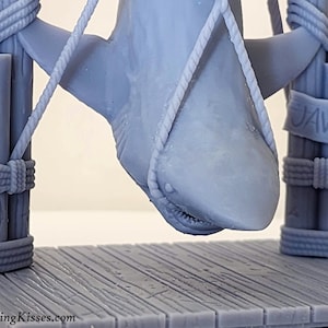 Pu&ograve; includere: Un modello 3D stampato in grigio di uno squalo appeso a una struttura in legno con una corda. Lo squalo &egrave; sospeso a testa in gi&ugrave; con la bocca aperta. La struttura ha un cartello con la scritta "JAWS".