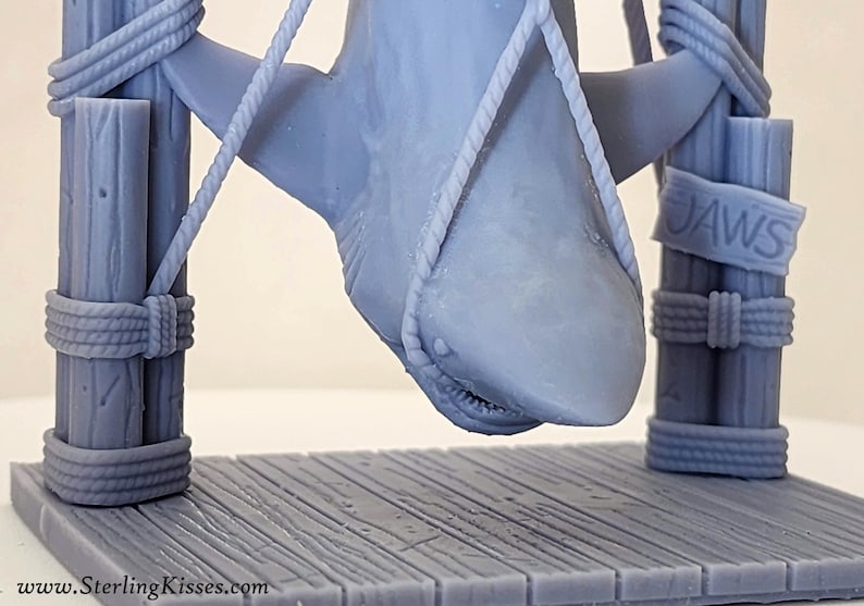 Pu&ograve; includere: Un modello 3D stampato in grigio di uno squalo appeso a una struttura in legno con una corda. Lo squalo &egrave; sospeso a testa in gi&ugrave; con la bocca aperta. La struttura ha un cartello con la scritta "JAWS".