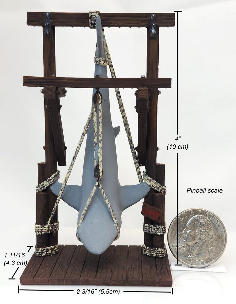 Pu&ograve; includere: Una statuetta di squalo grigia appesa a una cornice di legno con una corda. La cornice &egrave; alta 10 cm e larga 5,5 cm. La statuetta &egrave; etichettata "Jaws" ed &egrave; su una scala da flipper.
