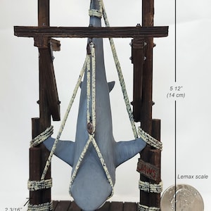 Pu&ograve; includere: Una statuetta di squalo grigio appesa a una struttura in legno con una corda. La struttura &egrave; etichettata "JAWS" e misura 14 cm di altezza e 7,8 cm di larghezza. Una moneta da 25 centesimi di dollaro americano &egrave; inclusa per la scala.