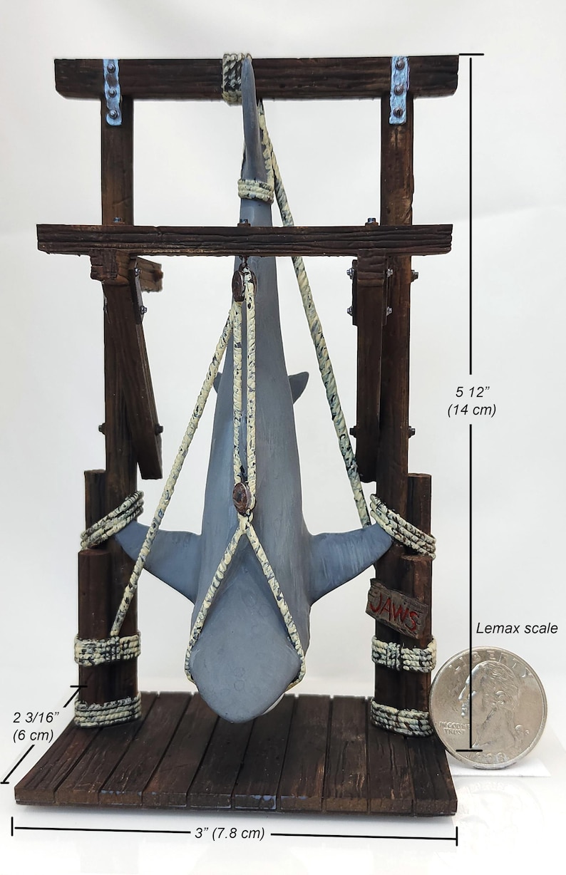 Pu&ograve; includere: Una statuetta di squalo grigio appesa a una struttura in legno con una corda. La struttura &egrave; etichettata "JAWS" e misura 14 cm di altezza e 7,8 cm di larghezza. Una moneta da 25 centesimi di dollaro americano &egrave; inclusa per la scala.